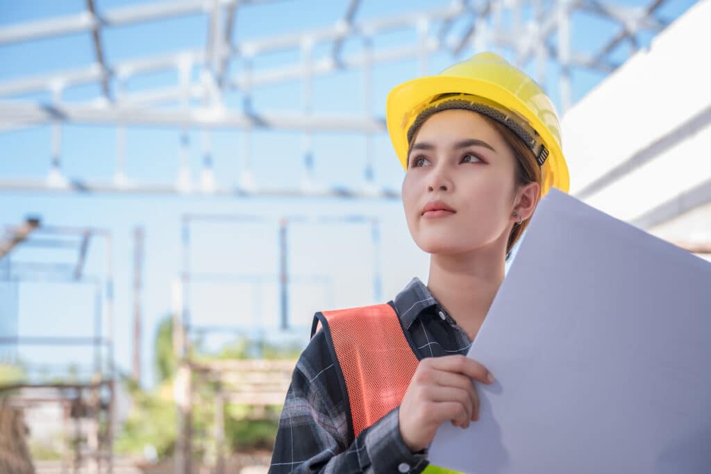 Asian woman civil engineer wear yellow safety hard hat standing and holding blueprint while checking infrastructure of building in construction site. Female architect supervisor working on a project.